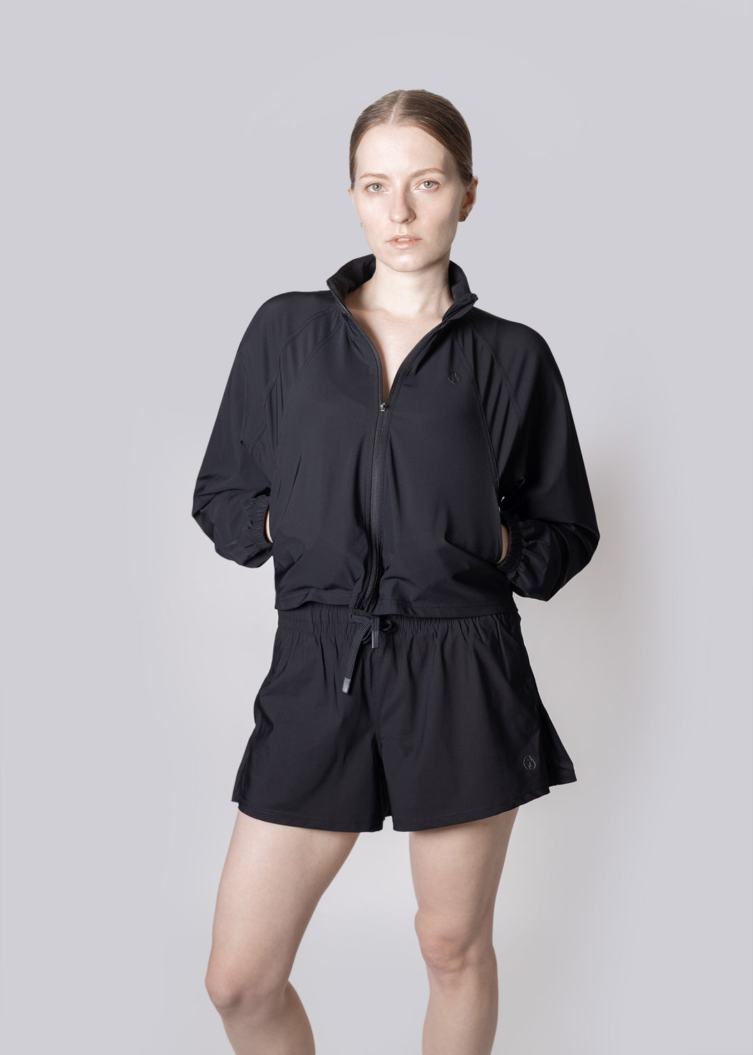 A person wearing a black outfit with a jacket and shorts on a plain background, with hands in the pockets